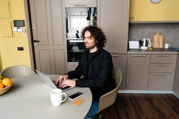Freelancer sorrindo digitando no laptop perto de smartphone com tela em branco, cartão de crédito e xícara de café — Fotografia de Stock