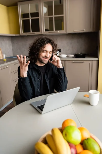 Selective focus of smiling freelancer gesturing while talking on smartphone near fresh fruits and coffee cup — Stock Photo