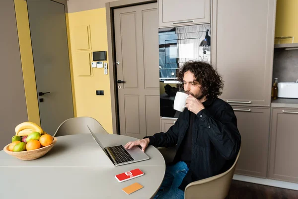 KYIV, UKRAINE - APRIL 25, 2020: handsome freelancer drinking coffee while using laptop near smartphone with Youtube app, credit card and fresh fruits — Stock Photo