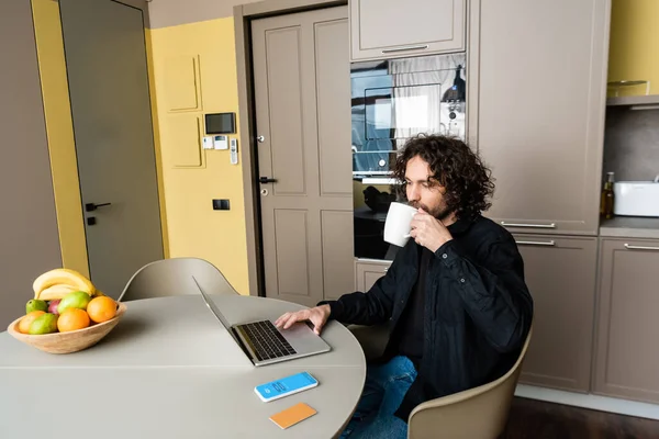 KYIV, UKRAINE - APRIL 25, 2020: handsome freelancer drinking coffee while using laptop near smartphone with Skype app, credit card and fresh fruits — Stock Photo