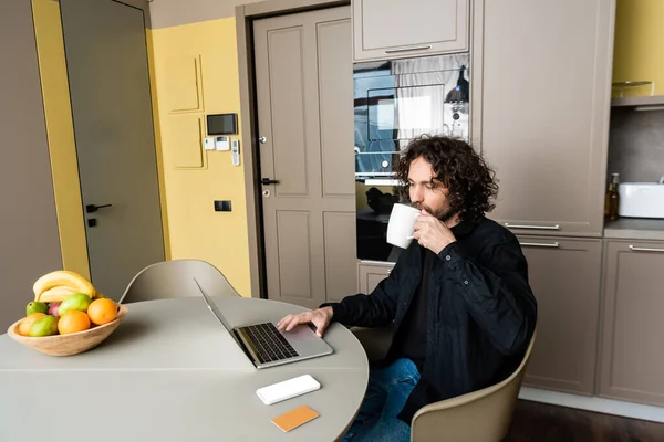 Guapo freelancer beber café mientras usa el ordenador portátil cerca de teléfono inteligente con pantalla blanca, tarjeta de crédito y frutas frescas - foto de stock