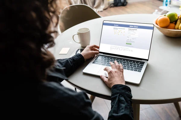 KYIV, UKRAINE - 25 AVRIL 2020 : vue arrière d'un pigiste utilisant un ordinateur portable avec Facebook à l'écran près d'une tasse de café, des notes collantes et des fruits — Photo de stock