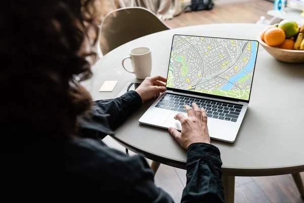 Vista posterior del freelancer utilizando el ordenador portátil con mapa en la pantalla cerca de la taza de café, notas adhesivas y frutas - foto de stock