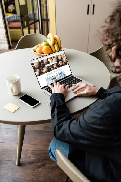 KYIV, UKRAINE - APRIL 25, 2020: back view of freelancer using laptop with Depositphotos website near smartphone, coffee cup and fresh fruits — Stock Photo