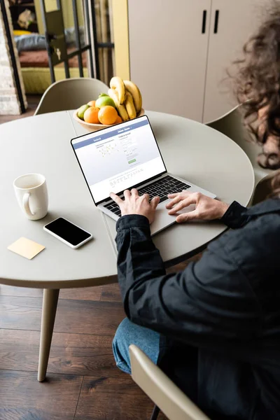 KYIV, UKRAINE - 25 de abril de 2020: visão traseira do freelancer usando laptop com Facebook na tela perto de smartphones, xícara de café, notas pegajosas e frutas — Fotografia de Stock