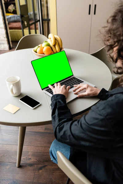 Back view of freelancer using laptop with green screen near smartphone, coffee cup and bowl with fresh fruits — Stock Photo