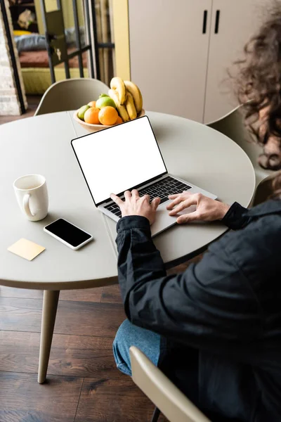 Vista posterior del freelancer utilizando el ordenador portátil con pantalla blanca cerca de teléfono inteligente, taza de café y tazón con frutas frescas - foto de stock