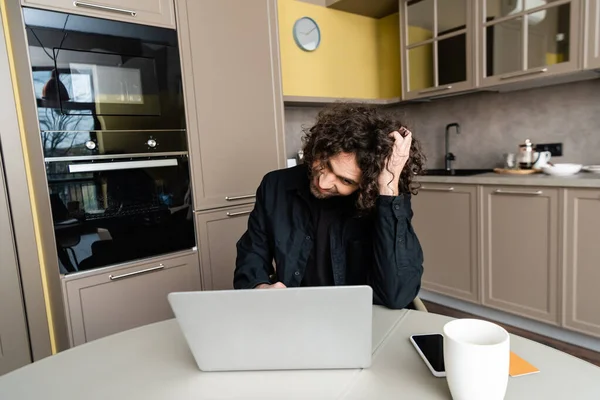 Reflexivo freelancer tocando la cabeza mientras se utiliza el ordenador portátil cerca de teléfono inteligente con pantalla en blanco y taza de café - foto de stock