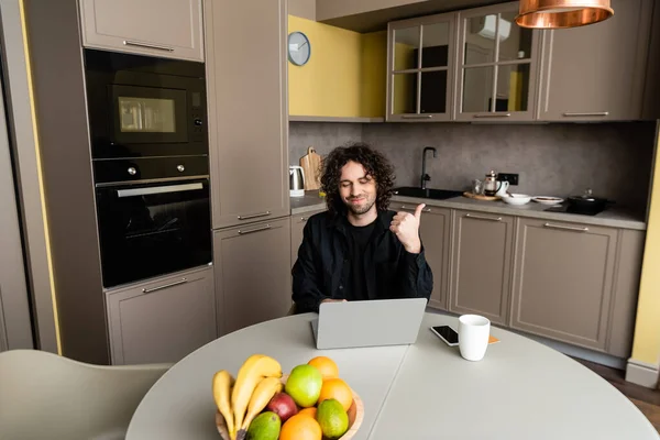 Freelancer sonriente mostrando el pulgar hacia arriba durante la videollamada en el portátil cerca del teléfono inteligente con pantalla en blanco y tazón con frutas frescas - foto de stock