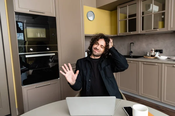 Smiling freelancer with closed eyes waving hand and touching hair during video chat on laptop near smartphone with blank screen — Stock Photo