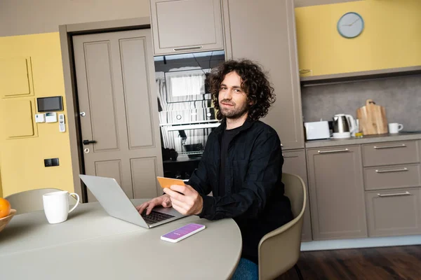 KYIV, UKRAINE - 25 AVRIL 2020 : beau freelance souriant à la caméra tout en tenant la carte de crédit et en utilisant un ordinateur portable près du smartphone avec l'application Instagram à l'écran — Photo de stock