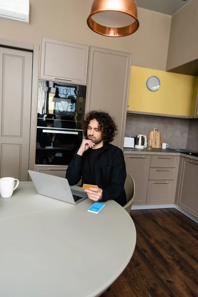 KYIV, UKRAINE - APRIL 25, 2020: thoughtful freelancer holding credit card while looking at laptop near smartphone with Skype app on screen — Stock Photo