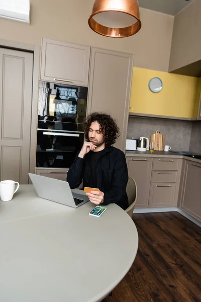 KYIV, UKRAINE - APRIL 25, 2020: thoughtful freelancer holding credit card while looking at laptop near Apple smarphone with homepage on screen — Stock Photo