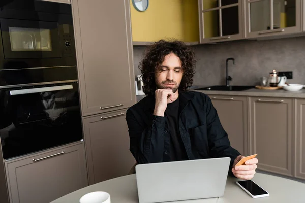 Thoughtful freelancer holding credit card while looking at laptop near smarphone with blank screen — Stock Photo