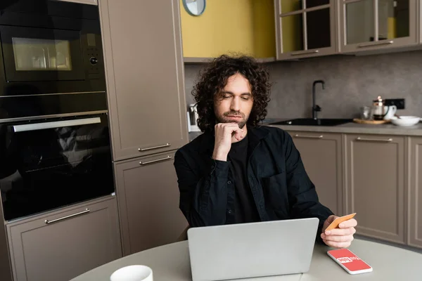 KYIV, UKRAINE - APRIL 25, 2020: pensive freelancer holding credit card near laptop and smartphone with Youtube app on screen — Stock Photo