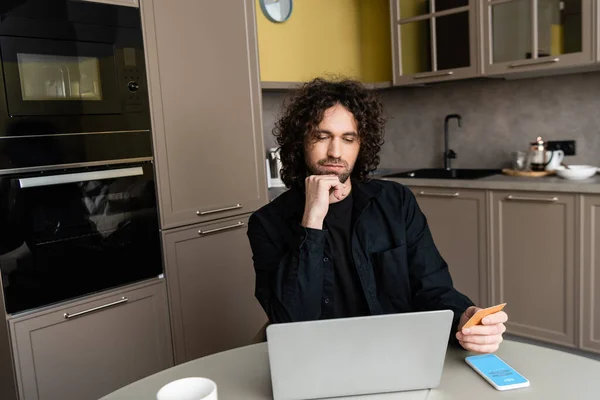 KYIV, UKRAINE - APRIL 25, 2020: pensive freelancer holding credit card near laptop and smartphone with Skype app on screen — Stock Photo