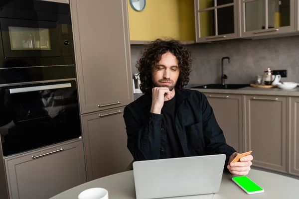 Pensativo freelancer celebración de la tarjeta de crédito cerca de la computadora portátil y el teléfono inteligente con pantalla verde - foto de stock