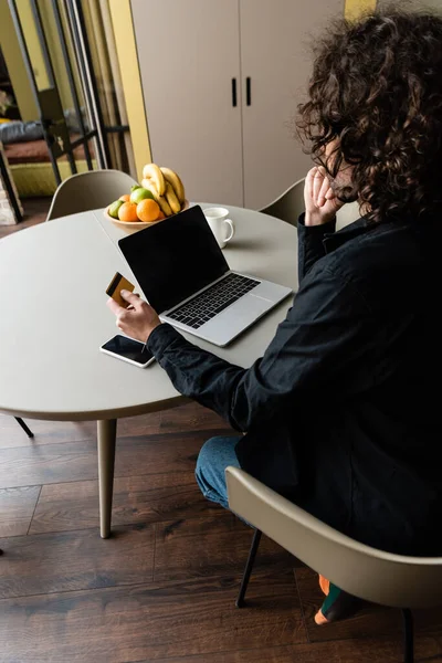 Back view of freelancer holding credit card while sitting at laptop with blank screen, smartphone and fruits — Stock Photo