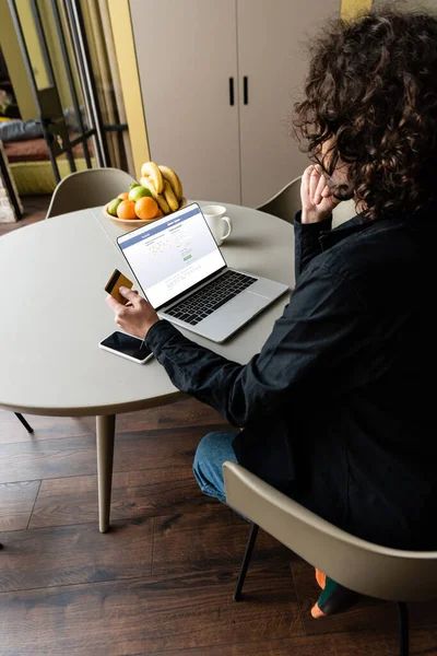 KYIV, UKRAINE - 25 de abril de 2020: visão traseira do freelancer segurando cartão de crédito perto do laptop com o site do Facebook, smartphone e frutas — Fotografia de Stock