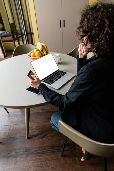 Back view of freelancer holding credit card while sitting at laptop with white screen, smartphone and fruits — Stock Photo