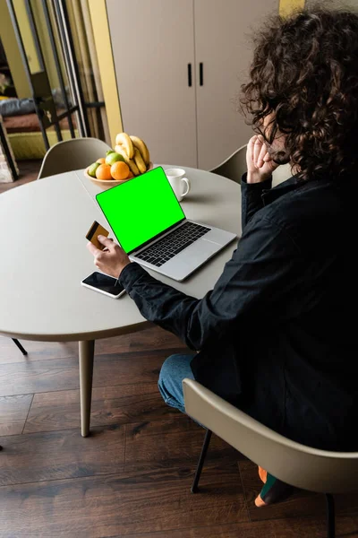 Vista trasera del freelancer que sostiene la tarjeta de crédito mientras está sentado en el ordenador portátil con pantalla verde, smartphone y frutas - foto de stock