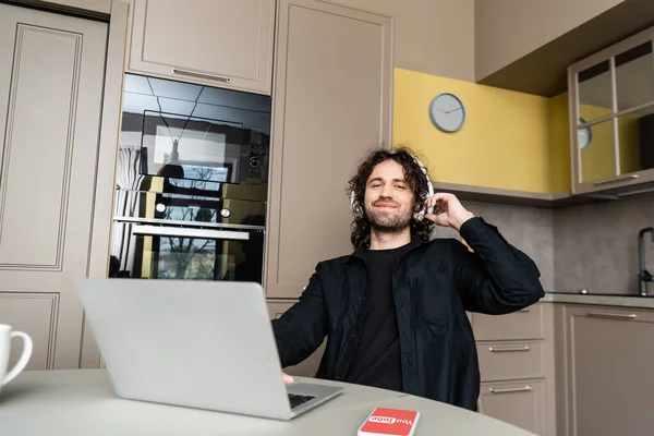 KYIV, UCRANIA - 25 DE ABRIL DE 2020: sonriendo freelance en auriculares inalámbricos mirando a la cámara cerca de la computadora portátil y el teléfono inteligente con la aplicación Youtube en la pantalla - foto de stock