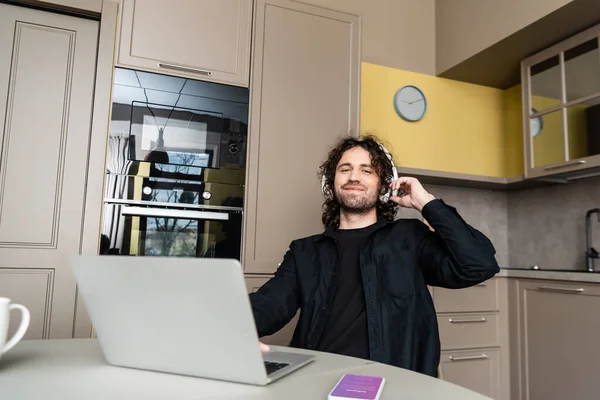 KYIV, UCRANIA - 25 DE ABRIL DE 2020: Enfoque selectivo del freelancer sonriente en auriculares con computadora portátil cerca de teléfono inteligente con aplicación instagram en la mesa de la cocina - foto de stock