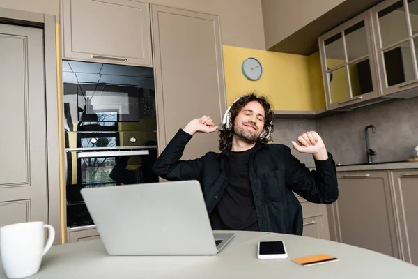 Выборочный фокус улыбающегося человека в наушниках, слушающего музыку возле гаджетов и кредитных карт на кухонном столе — стоковое фото