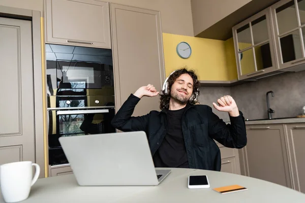 Enfoque selectivo del hombre sonriente en auriculares escuchando música cerca de gadgets y tarjeta de crédito en la mesa de la cocina - foto de stock