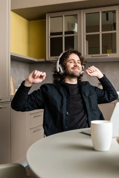 Enfoque selectivo de teletrabajador sonriente en auriculares sentados cerca de la computadora portátil y la taza en la mesa de la cocina - foto de stock