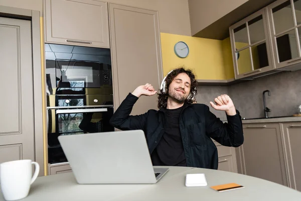 Enfoque selectivo del freelancer sonriente escuchando música en auriculares cerca de tarjeta de crédito, gadgets y taza en la mesa de la cocina - foto de stock