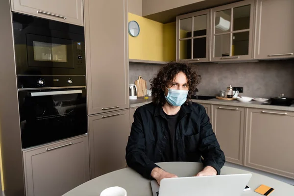Man in medical mask using laptop near smartphone and credit card on kitchen table — Stock Photo