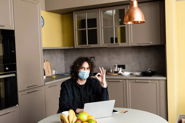 Hombre en máscara médica mostrando gesto ok cerca de gadgets y tarjeta de crédito en la mesa de la cocina - foto de stock