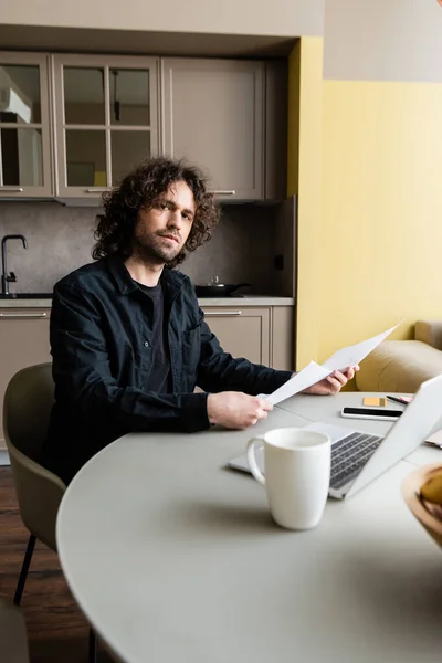 Selective focus of freelancer looking at camera while holding papers near gadgets, credit card and cup on table in kitchen — Stock Photo