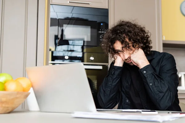 Selective focus of freelancer with hands near cheeks looking at camera while working with laptop and papers in kitchen — Stock Photo