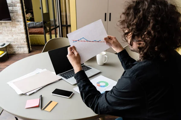 Rückansicht eines Freiberuflers, der Papier mit Diagrammen in der Nähe von Gadgets und Kreditkarte auf dem Küchentisch hält — Stockfoto