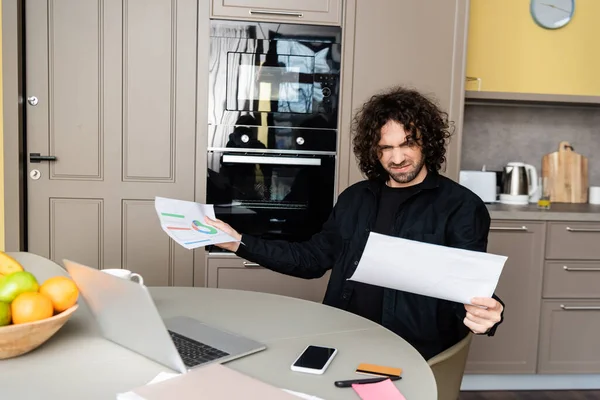 Selective focus of confused freelancer holding papers with charts near gadgets on kitchen table — Stock Photo