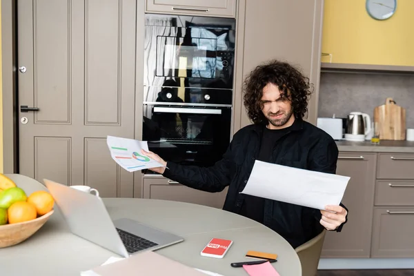 KIEW, UKRAINE - 25. April 2020: Selektiver Fokus eines verwirrten Telearbeiters, der Dokumente in der Nähe von Laptop, Smartphone mit Youtube-App und Kreditkarte auf dem Küchentisch hält — Stockfoto