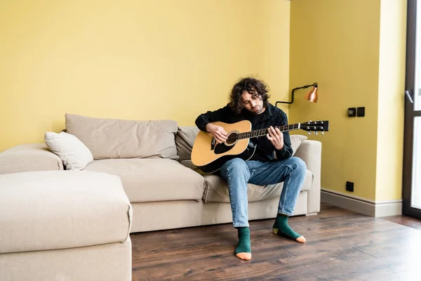 Curly homem tocando guitarra acústica enquanto sentado no sofá em casa — Stock Photo