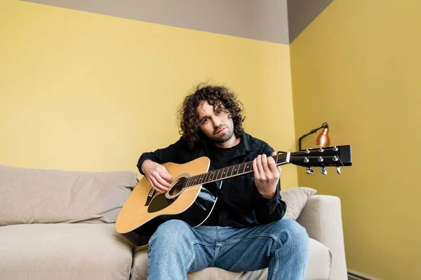 Bel homme regardant caméra tout en tenant guitare acoustique sur canapé — Photo de stock