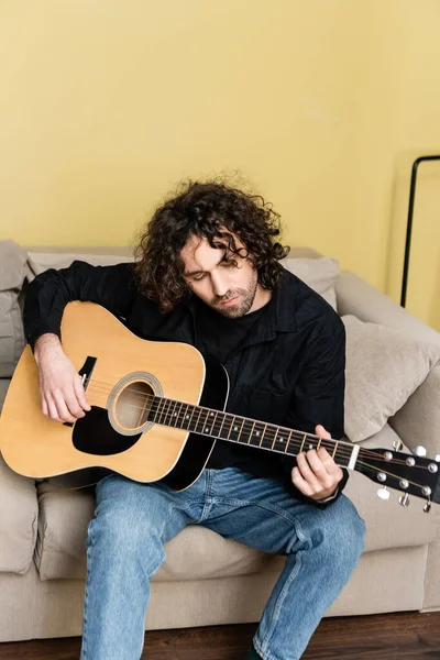 Beau bouclé homme jouant de la guitare acoustique sur le canapé dans le salon — Photo de stock