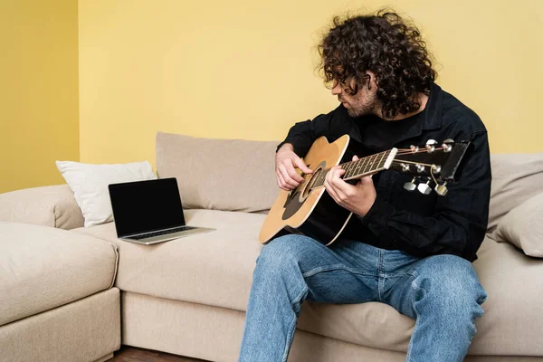 Hombre tocando la guitarra acústica durante el webinar en el sofá - foto de stock