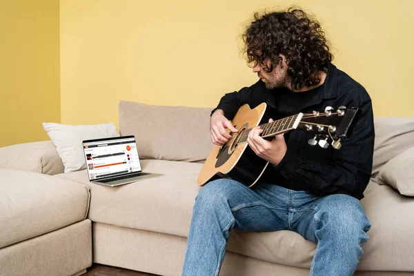 KYIV, UKRAINE - 25 de abril de 2020: Homem tocando guitarra acústica perto de laptop com site SoundCloud no sofá — Stock Photo