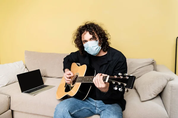 Человек в медицинской маске играет на акустической гитаре возле ноутбука на диване — стоковое фото