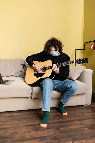 Людина в медичній масці грає на акустичній гітарі біля ноутбука під час вебінару вдома — стокове фото