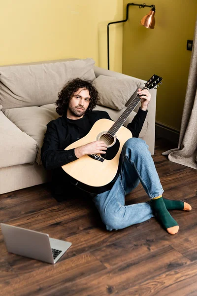 Красивий чоловік тримає акустичну гітару і дивиться на камеру біля ноутбука на підлозі — стокове фото