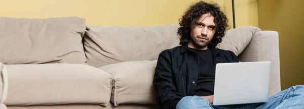 Panoramic orientation of handsome man looking at camera while using laptop on floor in living room — Stock Photo