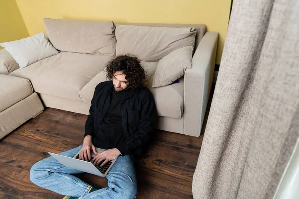 Selective focus of curly man using laptop on floor at home — Stock Photo
