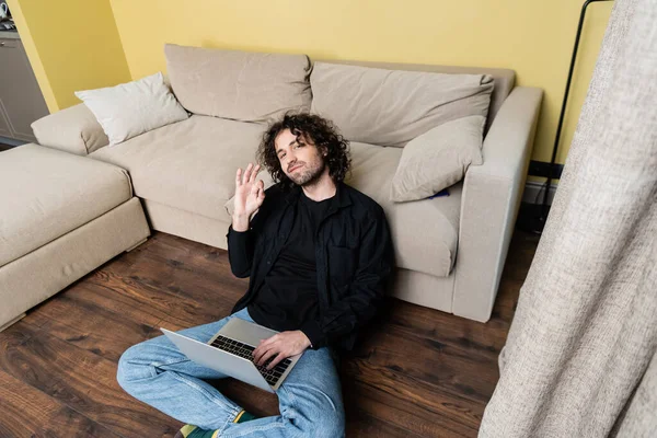 Foco seletivo de homem bonito mostrando gesto ok ao usar laptop no chão — Fotografia de Stock