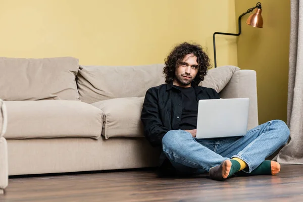 Baixo ângulo de visão de homem bonito encaracolado olhando para a câmera ao usar laptop no chão na sala de estar — Fotografia de Stock